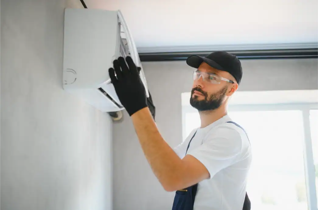 A technician from a professional AC company, wearing protective glasses and gloves, is inspecting or repairing a wall-mounted air conditioner indoors.