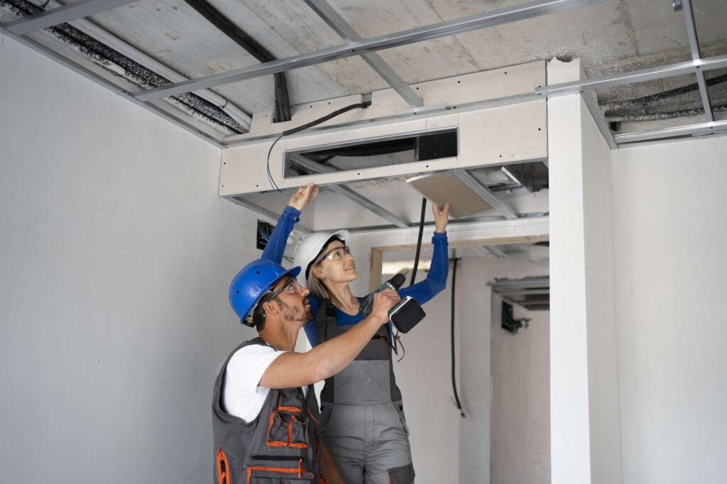 Two construction workers in safety gear inspect and discuss the installation of an overhead ceiling panel, ensuring the space meets standards for future commercial AC services in the unfinished room.