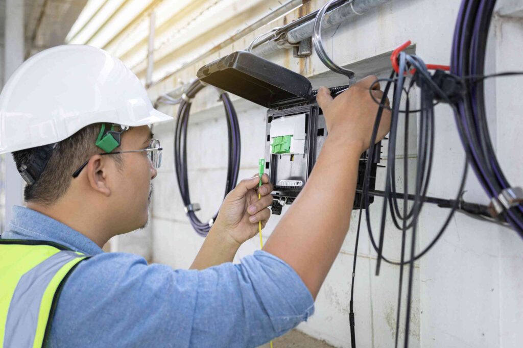 An electrical contractor in a hard hat and safety vest installs or repairs cables in a wall-mounted electrical or fiber optic box.