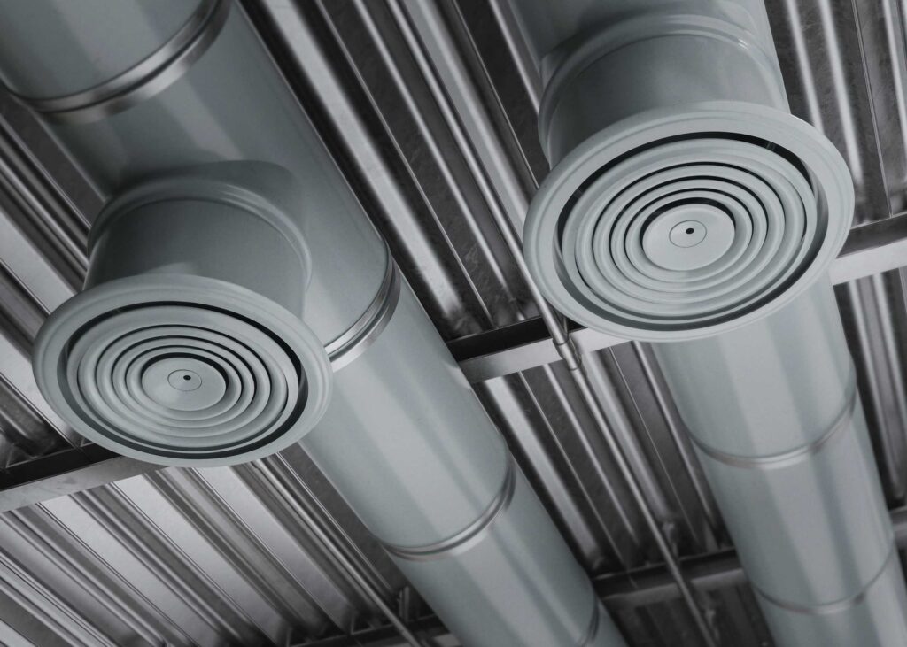 Two large circular air vents attached to metal ventilation ducts, recently serviced with duct cleaning, are installed on a corrugated metal ceiling.