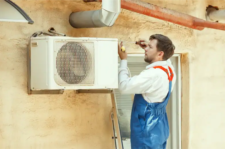 A technician in blue coveralls repairs or installs an outdoor air conditioning unit on a building's exterior wall, addressing issues like warm air from AC or other signs your AC needs repair.