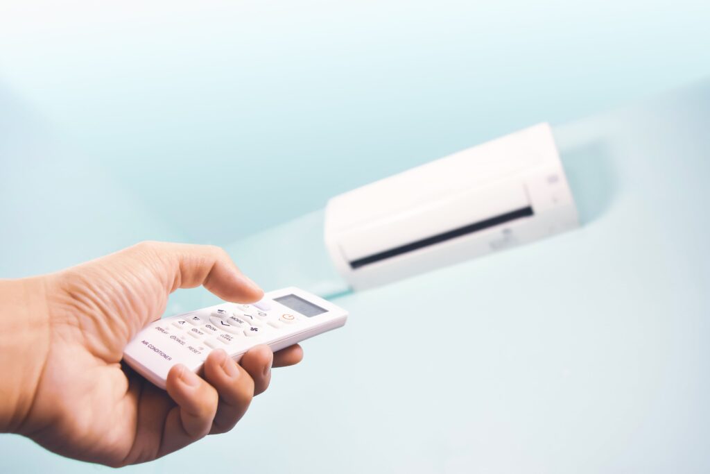 A hand holding a remote control pointed at a wall-mounted air conditioner unit against a light blue background, showcasing the comfort and efficiency provided by professional HVAC services Miami.